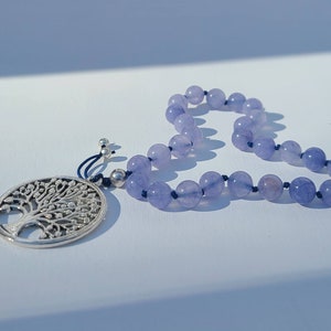May include: A lavender coloured beaded necklace with a silver tree of life pendant. The necklace is made of round beads and is strung on a dark blue cord.