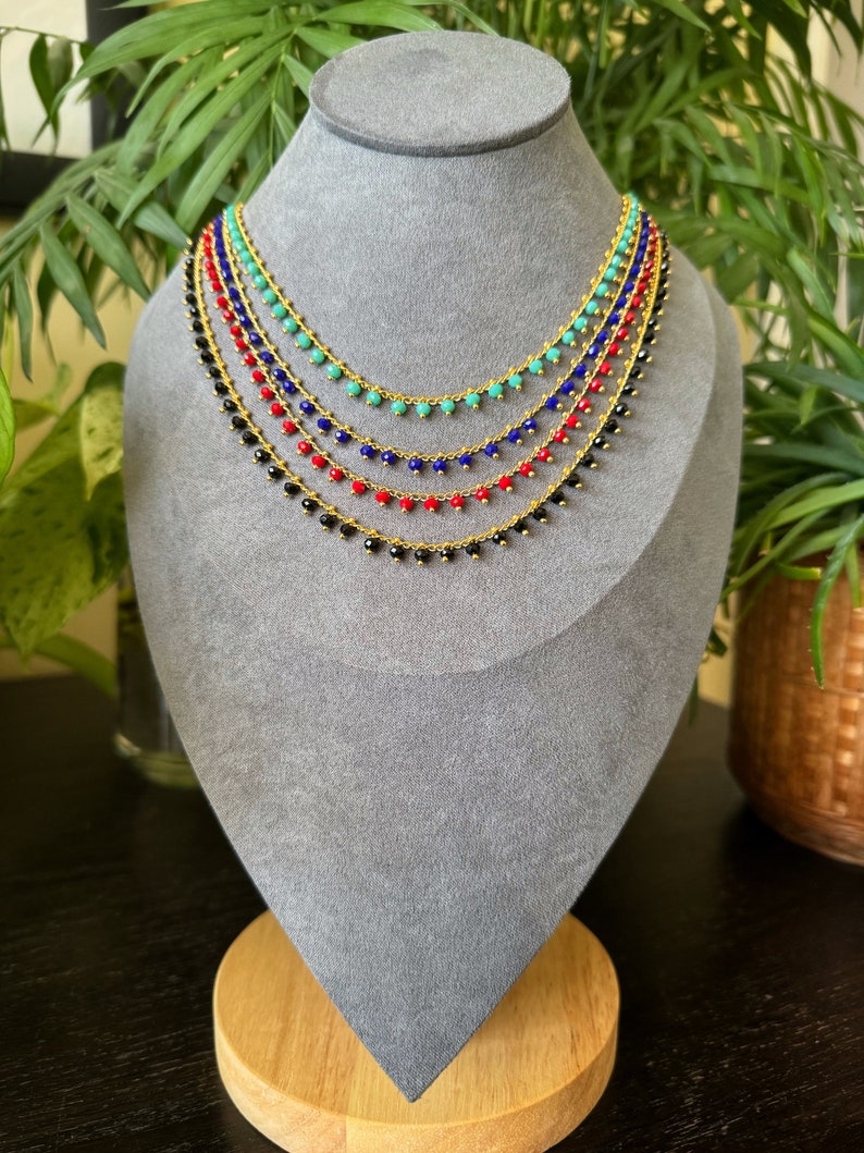Peut inclure: Cinq colliers superpos&eacute;s avec des cha&icirc;nes dor&eacute;es et de petites perles. Les perles sont noires, rouges, bleues et turquoise.