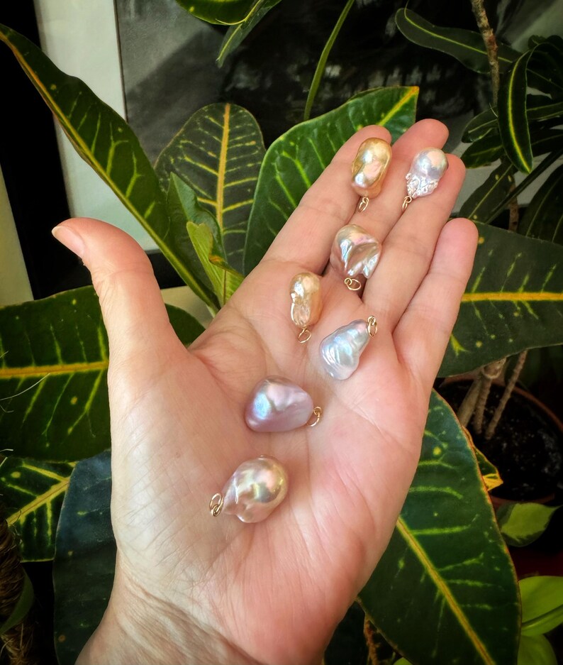 以下が含まれることがあります： 金色のワイヤーのループが付いた、形が不規則な虹色の真珠のコレクション。真珠の色は白、ピンク、金など様々で、緑の葉を背景に展示されています。