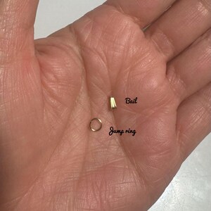 以下が含まれることがあります： 金色のジャンプリングとベイルのクローズアップ。ジャンプリングは、小さな円形のジュエリーパーツです。ベイルは、小さな長方形のジュエリーパーツです。「Bail」と「Jump ring」の文字が黒で書かれています。