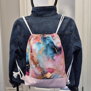 Peut inclure: Un sac à dos à cordon avec un motif marbré abstrait multicolore dans des tons de bleu, rose et doré, doté d'une base rose clair unie, d'une fermeture éclair dorée verticale sur le côté gauche et de épaisses cordes blanches tressées.