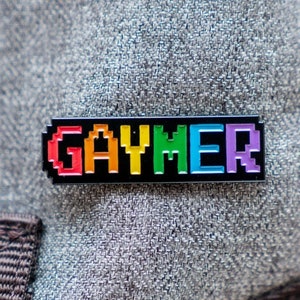 Puede incluir: Un pin de esmalte negro con la palabra "GAYMER" en letras pixeladas de arcoíris.