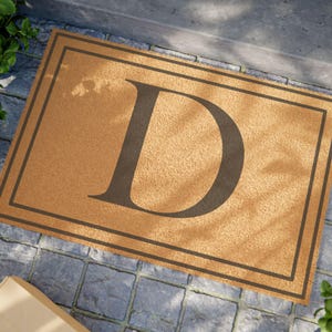 May include: A rectangular doormat with a large, dark brown letter "D" in the center. The mat is a light brown color with a darker brown border. The doormat is placed on a gray stone surface.
