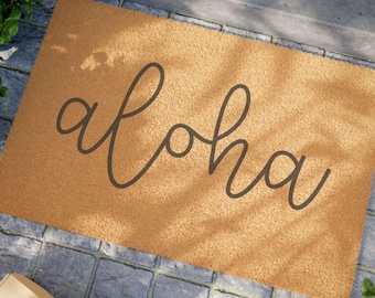 May include: A rectangular doormat with the word "aloha" in a dark gray script. The mat is a light brown color and sits on a gray stone surface. Green plants are visible in the corners.