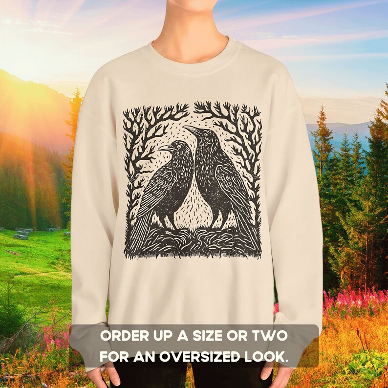 May include: A beige sweatshirt with a black and white illustration of two crows perched on a branch. The illustration is framed by a square of stylized branches. The text "ORDER UP A SIZE OR TWO FOR AN OVERSIZED LOOK." is printed below the illustration.