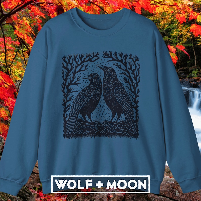 May include: A dark blue sweatshirt with a black and white illustration of two crows perched on branches. The illustration is framed by a square border. The text "WOLF + MOON" is printed in white on the bottom of the sweatshirt.