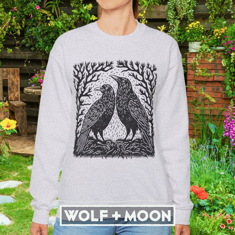 May include: A gray sweatshirt with a black and white illustration of two crows facing each other in a forest setting. The text "WOLF + MOON" is printed on the bottom of the sweatshirt.