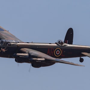 Pode incluir: Um avião bombardeiro Avro Lancaster vintage, pintado em camuflagem verde escuro e marrom com marcas vermelhas, brancas e azuis, incluindo um círculo e o texto "AROL" no lado. O avião está em voo contra um céu azul claro.