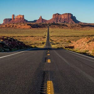 Könnte beinhalten: Eine lange, gerade Asphaltstraße erstreckt sich zum Horizont, flankiert von Wüstenvegetation und roten Felsformationen unter einem klaren blauen Himmel. Gelbe Fahrbahnmarkierungen weisen den Weg und erzeugen eine Tiefenwirkung in dieser Landschaft.