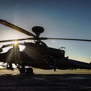 May include: A black military helicopter with a large rotor blade, silhouetted against a bright orange sunset. The helicopter is on a runway with a blurry background.