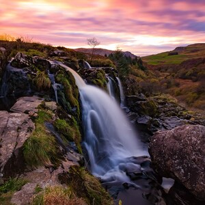 Può includere: Una cascata scende a cascata su rocce ricoperte di muschio, con un vibrante tramonto rosa e viola sullo sfondo. L'acqua scorre dolcemente, creando un senso di tranquillità.