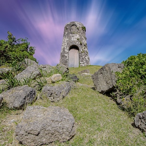 May include: A stone windmill with a wooden door stands on a grassy hill with scattered rocks. The sky is a vibrant blue with streaks of pink and purple clouds.