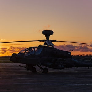 May include: Silhouette of a military helicopter on a runway at sunset. The helicopter is dark grey and has a large rotor on top. The sun is setting in the background, casting a warm glow on the sky.