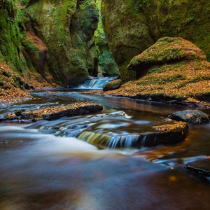 May include: A stream flows through a rocky, moss-covered gorge. The water is clear and blue, and the rocks are covered in fallen leaves. The stream is surrounded by lush green trees.