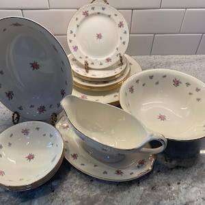 Puede incluir: Una colección de vajilla de porcelana vintage con un estampado floral. El juego incluye platos, cuencos y fuentes para servir, todos con una base de color crema, ribete dorado y delicados detalles florales rosas y azules. La disposición muestra la variedad de piezas.