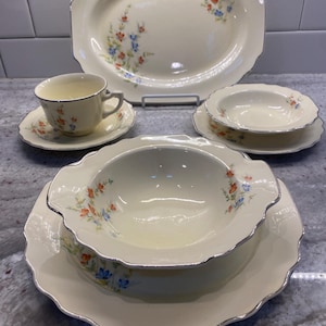 May include: A cream-colored dinnerware set featuring a scalloped edge with silver trim and a delicate floral pattern of orange and blue flowers. The collection includes a large oval platter, a teacup with a saucer, and two shallow bowls.