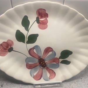 Może przedstawiać: Kremowy ceramiczny talerz z falistym brzegiem, zdobiony ręcznie malowanymi kwiatami w odcieniach czerwieni, błękitu i zieleni. Kwiatowy wzór jest umieszczony centralnie na talerzu, z zielonymi liśćmi i łodygami.
