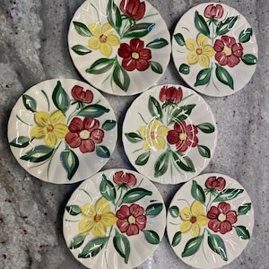 Puede incluir: Juego de seis platos de cerámica blanca, cada uno adornado con un diseño floral pintado a mano. Las flores presentan pétalos amarillos y burdeos con hojas verdes. Los platos están dispuestos sobre una superficie gris moteada.