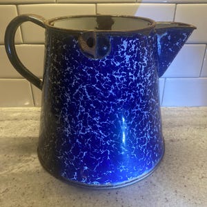 Puede incluir: Jarra de esmalte azul y blanco con pico vertedor y asa. La jarra presenta un patrón moteado y un borde oscuro. El asa y el borde muestran signos de desgaste, lo que añade encanto vintage. Un artículo de cocina clásico.