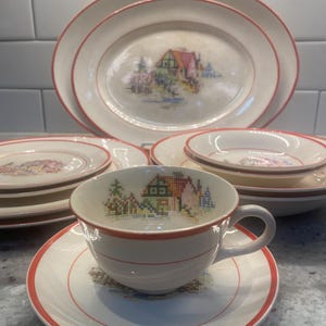 Könnte beinhalten: Ein cremefarbenes Vintage-Teeservice mit rotem Rand und Kreuzstichmuster eines Hauses und Bäumen. Das Set umfasst ovale Platten und Teller mit passenden Designs, perfekt für eine Teeparty oder zur Präsentation.