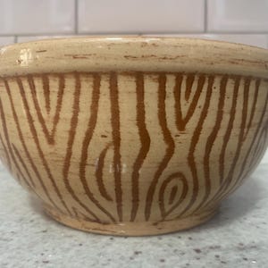 Puede incluir: Un cuenco de cerámica beige con un patrón de vetas de madera en marrón. El cuenco tiene forma redondeada y una abertura ancha. El diseño de vetas de madera cubre el exterior del cuenco, creando una estética rústica. Este cuenco es adecuado para servir alimentos.