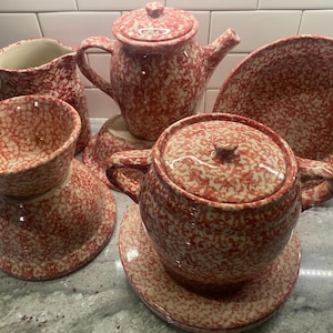 May include: A collection of red and cream-colored speckled ceramic kitchenware, including a teapot, a creamer, a bowl, a covered serving dish with a saucer, and a plate. The pieces are arranged on a light-colored surface.