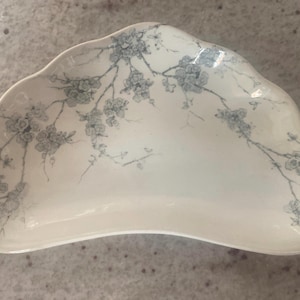 May include: A white, scalloped-edge serving dish with a delicate floral pattern in shades of blue. The dish has an unusual, asymmetrical shape, ideal for serving appetizers or small dishes. The floral design adds a touch of elegance.