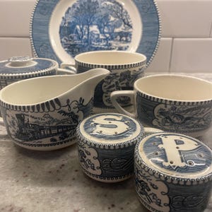 Puede incluir: Una colección de vajilla de cerámica azul y blanca, que incluye platos, tazas, una crema y saleros y pimenteros. Las piezas presentan un diseño escénico con un pueblo y un tren. Los saleros y pimenteros están etiquetados con las letras "S" y "P".