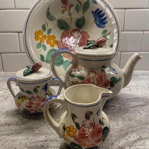 Op de afbeelding: Een vintage keramische theeset met een grote serveerschaal, een theepot, een melkkan en twee kleine suikerpotten met deksel. De set is versierd met handgeschilderde bloemmotieven in roze, geel en blauw, met groene bladeren. De randen zijn blauw afgewerkt.
