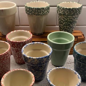 Puede incluir: Una colección de macetas de cerámica en varios colores y patrones. Las macetas incluyen crema, rojo, azul y verde, con diseños moteados y sólidos. Las macetas están dispuestas sobre una superficie de madera.