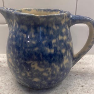 May include: A blue and cream-colored ceramic pitcher with a rounded body and a curved handle. The pitcher has a speckled pattern and a spout for pouring. The handle and rim are cream-colored.