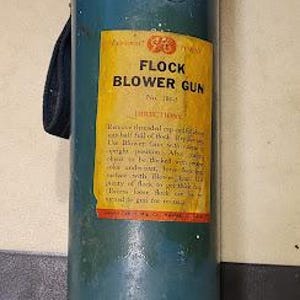 May include: Vintage teal Flock Blower Gun with a yellow label and red accents. The label reads "Flock Blower Gun" with instructions. The gun has a handle and a red base. The metal shows signs of wear.