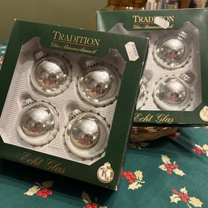 May include: Two boxes of silver glass Christmas baubles. Each box contains four baubles. The boxes are dark green with the words "Tradition Glas-Baumschmuck" and "Echt Glas" printed on them. The baubles are reflective.