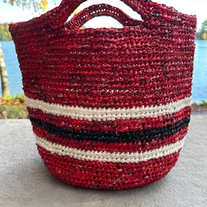 May include: A handmade red tote bag with two handles. The bag features horizontal stripes of white and black near the base. The bag is made of woven material and is suitable for carrying items. The background shows a lake and trees.