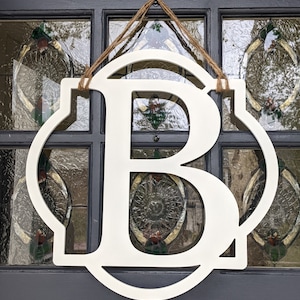 May include: A white, circular, flat-cut wooden sign featuring a large, centered capital letter B. The sign is suspended by a thin, brown, twisted jute rope attached to the top, creating a decorative hanging display for a door or wall.