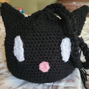 May include: Black crocheted cat-shaped drawstring bag with white eyes and a pink nose.
