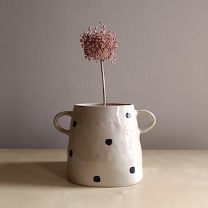 Könnte beinhalten: Eine cremefarbene Keramikvase mit zwei seitlichen Henkeln und einem handgemalten schwarzen Punktemuster. Die Vase hat eine unebene, organische Textur und enthält einen einzelnen getrockneten rosa Blumenstiel in der Öffnung.