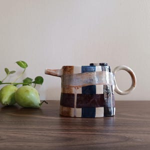 Könnte beinhalten: Eine Keramik-Teekanne mit zylindrischem Körper und einem handgemalten Karomuster in Dunkelbraun, Marineblau und Creme. Die Teekanne hat einen kurzen Ausguss, einen runden Henkel und einen flachen Deckel mit sichtbaren Glasurtropfen auf der Oberfläche.