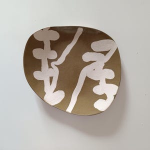 Könnte beinhalten: Ein handgefertigter, olivgrüner Keramikteller mit unregelmäßiger Form. Der Teller hat ein weißes, abstraktes Design, das auf seine Oberfläche gemalt wurde. Das Design besteht aus organischen Formen und Linien und schafft eine einzigartige, künstlerische Ästhetik.