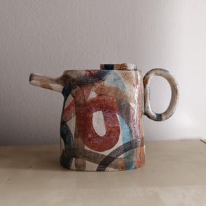 Op de afbeelding: Een theepot van keramiek met een uniek abstract ontwerp in tinten bruin, rood, blauw en wit. De theepot heeft een rond lichaam en een tuit aan de ene kant. De handgreep bevindt zich aan de andere kant van de tuit.