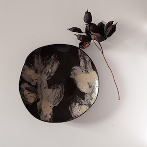 Può includere: Un piatto rotondo in ceramica nera con disegni astratti a pennellate beige e grigie. Uno stelo di pianta secca, marrone scuro, con baccelli di semi è posizionato a destra del piatto, su sfondo bianco. Il piatto ha una finitura lucida.
