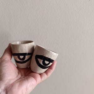 Può includere: Due piccole tazze in ceramica macchiettata con base color crema. Ogni tazza presenta un audace disegno di occhi neri. Le tazze sono tenute in mano, mostrando le loro dimensioni e forma. Il design è minimalista e moderno.