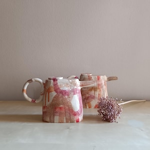 Op de afbeelding: Twee keramische theepotten met een glanzende afwerking, voorzien van abstracte, handgeschilderde patronen in roze, rode en bruine tinten. Elke theepot heeft een cirkelvormig handvat, een korte tuit en een plat deksel. Een gedroogde bloem ligt ernaast.