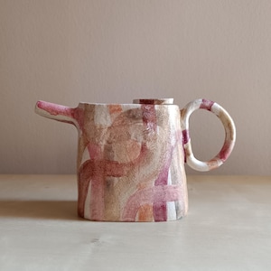 May include: A ceramic teapot with a lid, spout, and handle. The teapot features a watercolour design in pink, beige, and white. The handle and spout are a solid pink colour.