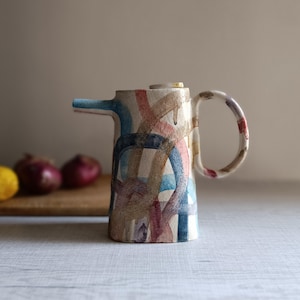 Könnte beinhalten: Ein Keramikkrug mit Ausguss und Henkel, verziert mit bunten abstrakten Pinselstrichen in Blau, Rosa und Braun. Der Krug steht auf einer weißen Oberfläche, mit einem Holzschneidebrett und Gemüse im Hintergrund.