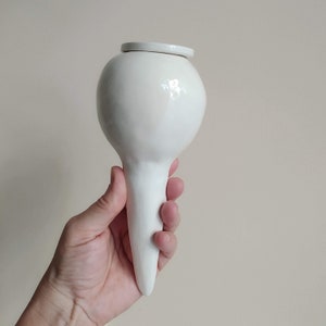 Può includere: Un oggetto in ceramica bianca con una parte superiore arrotondata e una parte inferiore lunga e appuntita. L'oggetto è tenuto in una mano.