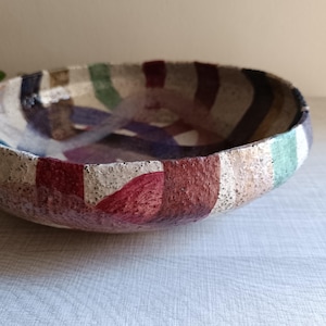 Pode incluir: Uma tigela de cerâmica redonda e rasa com uma base texturizada e salpicada em tom branco sujo. O exterior e o interior são decorados com largas faixas verticais pintadas em tons de vermelho escuro, castanho, verde floresta e azul marinho.