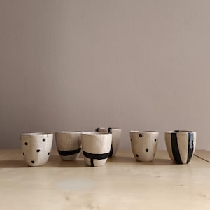 Può includere: Una collezione di sei piccole tazze in ceramica bianco sporco con disegni neri dipinti. I disegni includono pois neri, strisce orizzontali e verticali. Le tazze sono disposte su una superficie di legno chiaro.