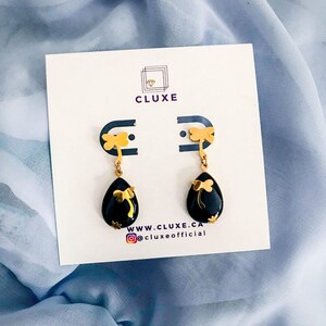 Peut inclure: Une paire de boucles d'oreilles pendantes dorées avec des pierres noires en forme de larme et un motif de libellule dorée. Les boucles d'oreilles sont sur une carte blanche avec le texte "CLUXE" et "WWW.CLUXE.CA @cluxeofficial".