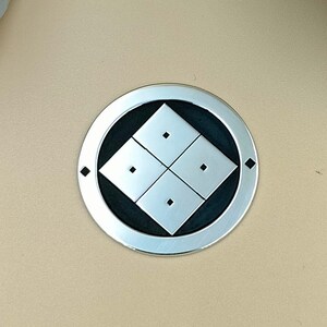 Pode incluir: Um emblema prateado com um design geométrico em preto e branco, apresentando quatro formas de diamante e quatro pequenos pontos. O emblema está sobre um fundo bege, com várias pedras na parte superior da imagem.